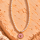 A white pearl necklace with a gold pendant featuring a pink enamel background and a gold starfish, displayed on a textured peach-colored stone.